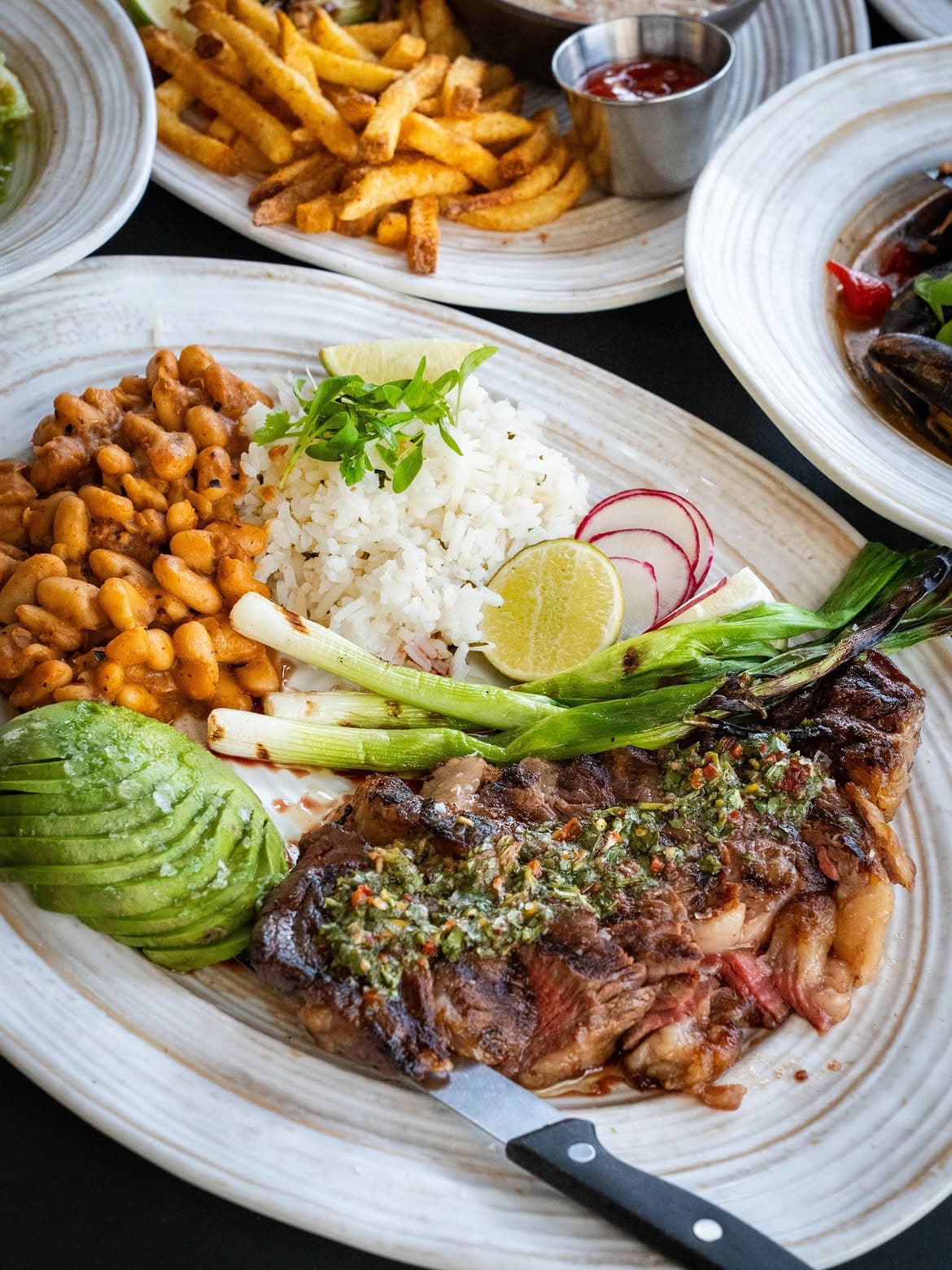 High Marea ribeye plate with chimichurri, avocado, rice, beans and grilled onions.